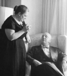 black and white photo of woman standing speaking with bald man seated.