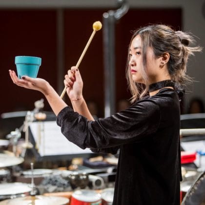 A percussionist prepares to strike a small blue pot with a yellow mallet, in a performance of the MSM Percussion Ensemble