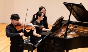 A violist plays while accompanied by a pianist, assisted by a page turner, during a concert at Manhattan School of Music's Greenfield Hall